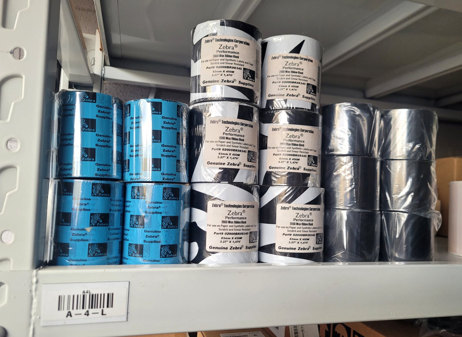 A printed bin location label reading A-4-L, on a magnetic strip fixed to a
                  warehouse racking beam, with rolls of Zebra printer ribbon stacked on the shelf
                  above it