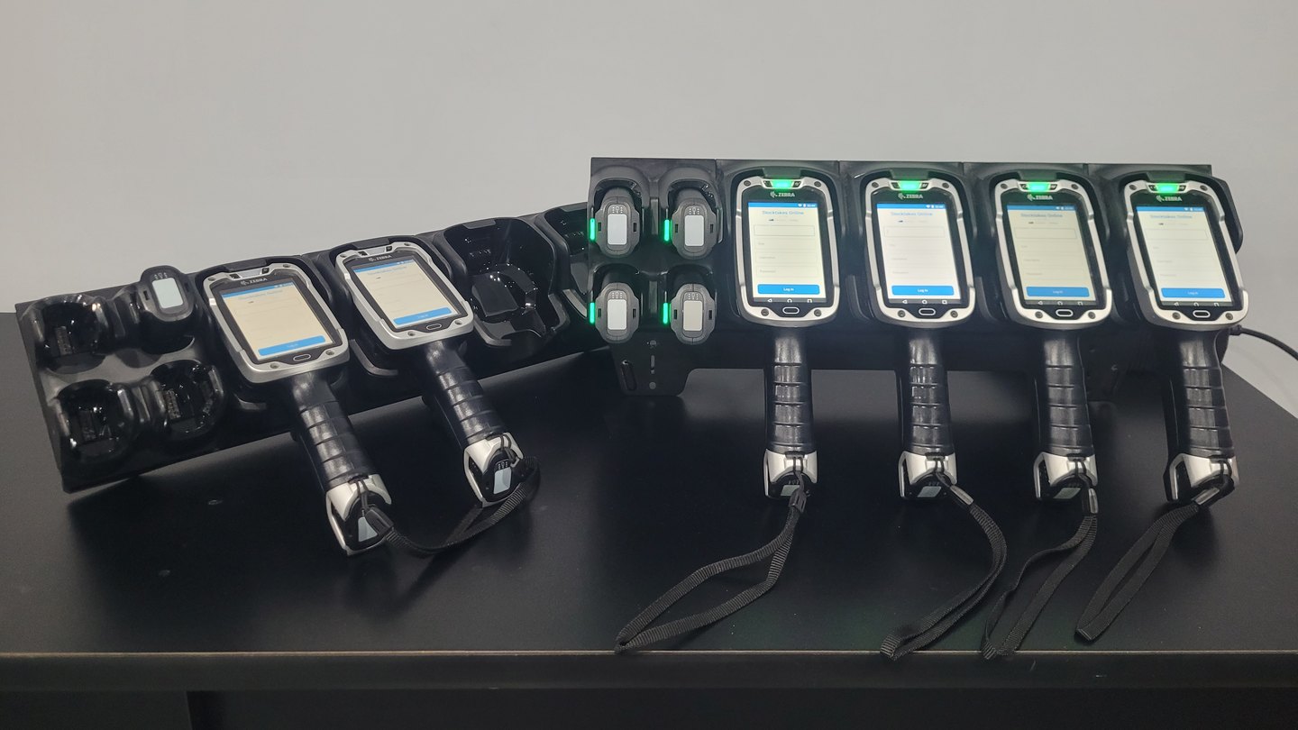 Six Zebra TC8000 scanners standing in two charging cradles on a desk, each
                  showing the Stocktakes Online sign-in screen, with spare batteries charging
                  in the bays beside them